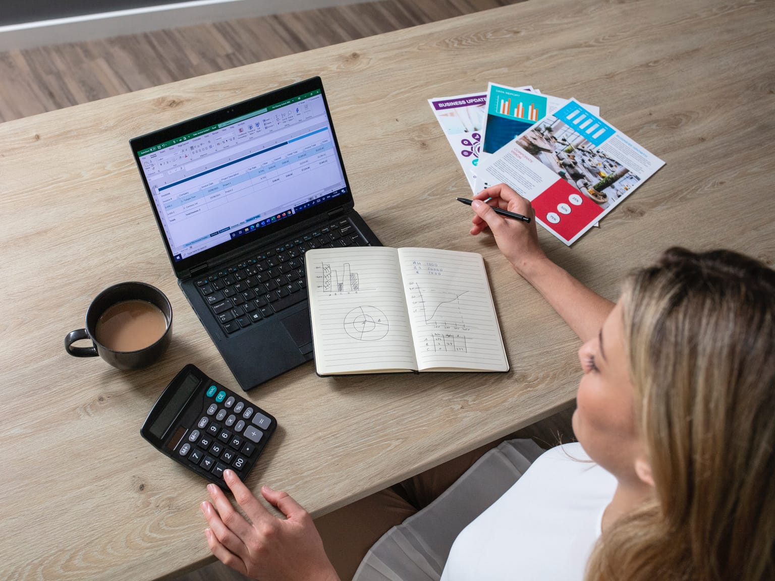 A female business owner calculating costs while sat at a desk with a notebook, calculator and laptop computer