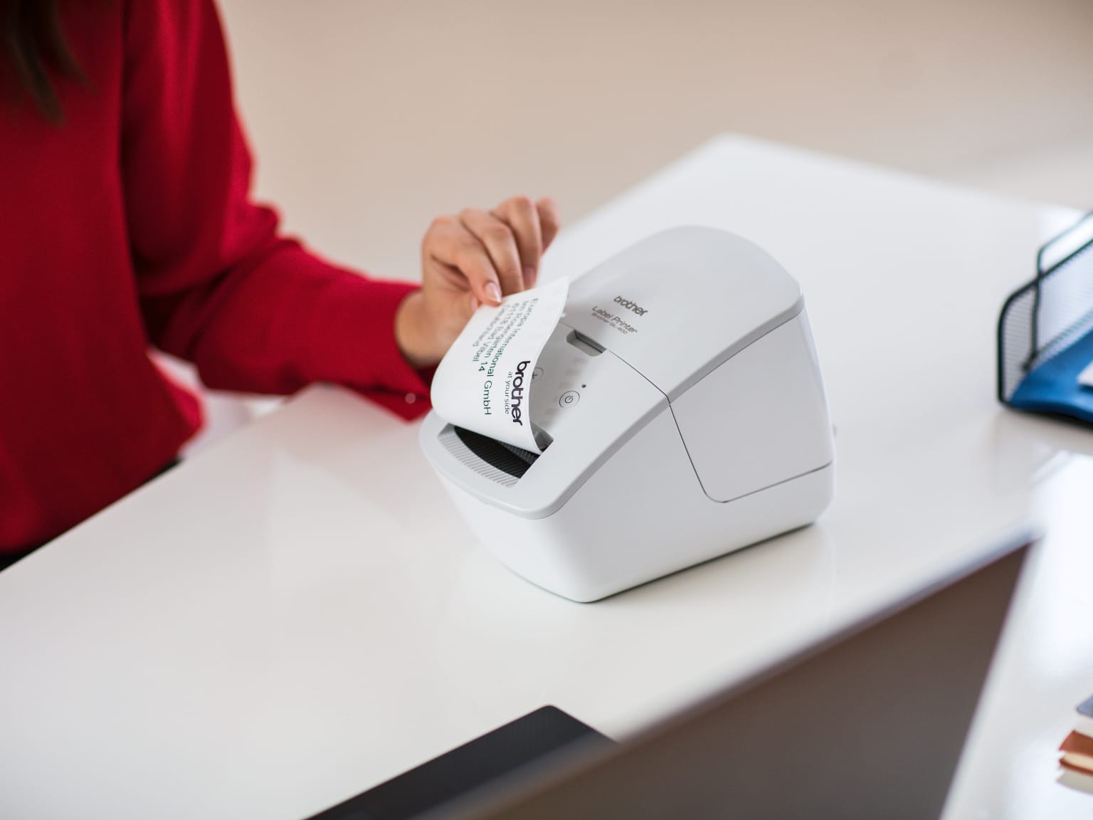 A lady wearing a red top removing a printed address label from a Brother thermal label printer which is on a desk in an office environment