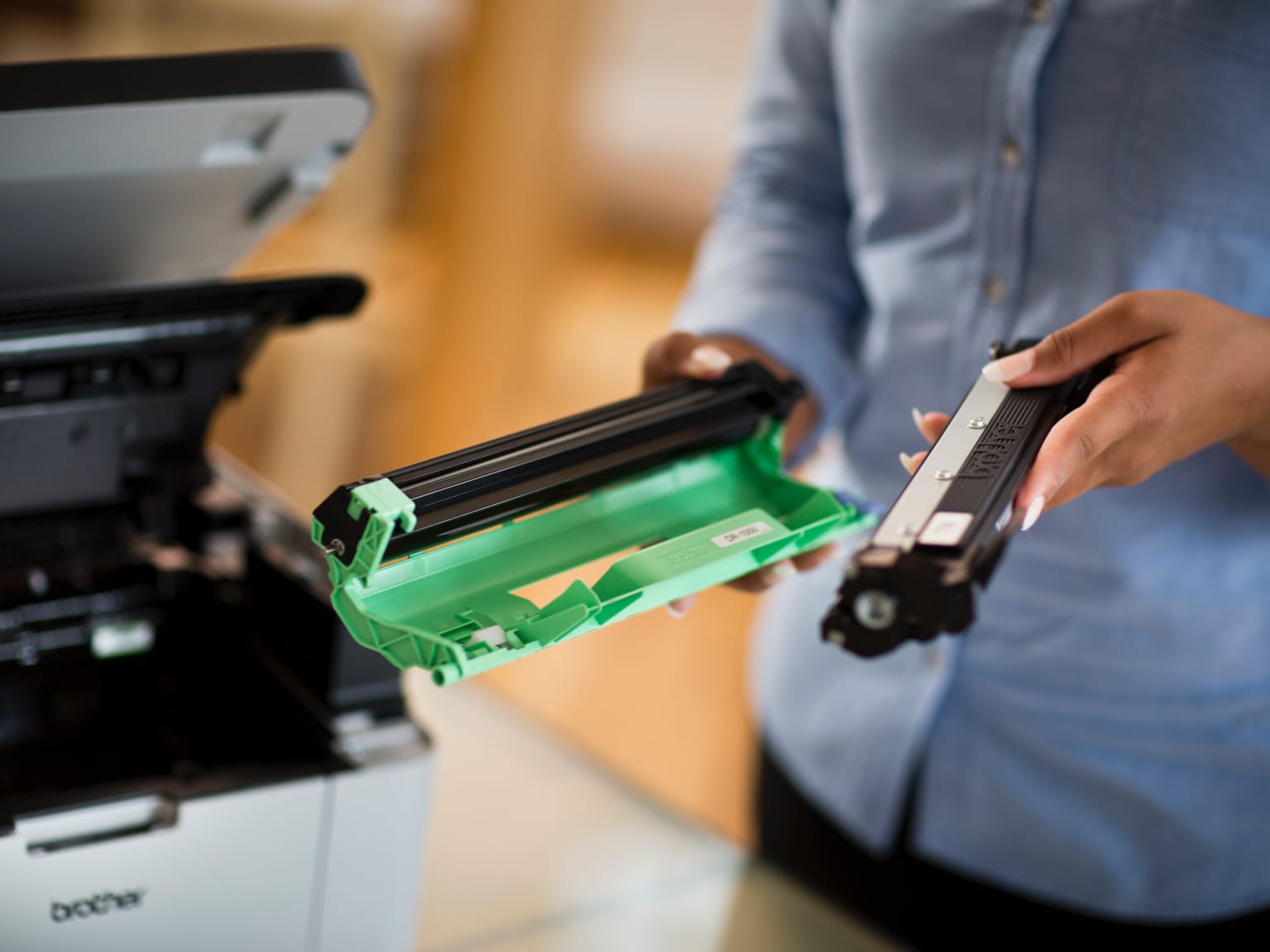 A lady holding two parts of a toner cartridge while standing next to an open laser printer