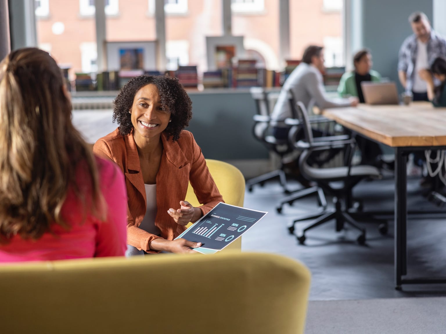 Two female colleagues discussing printed statistics in an office environment with a meeting around a desk in the background