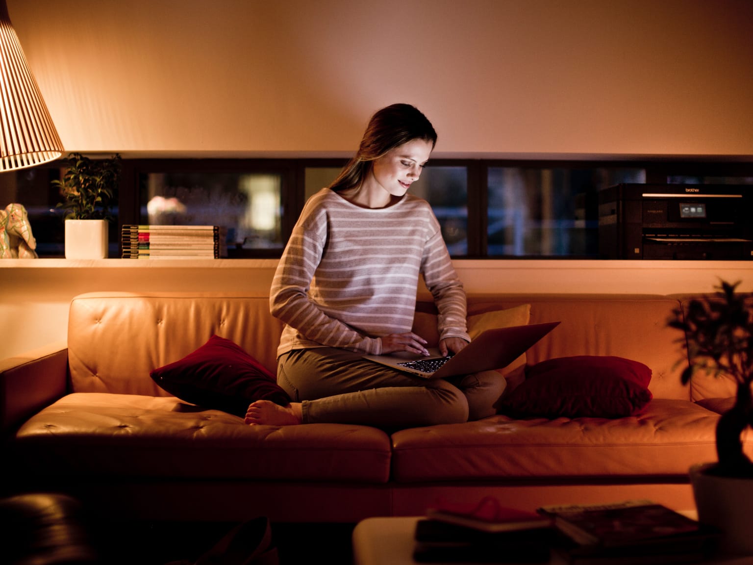 A woman printing an email from a notebook computer while relaxing on a brown leather sofa in a softly lit home living room