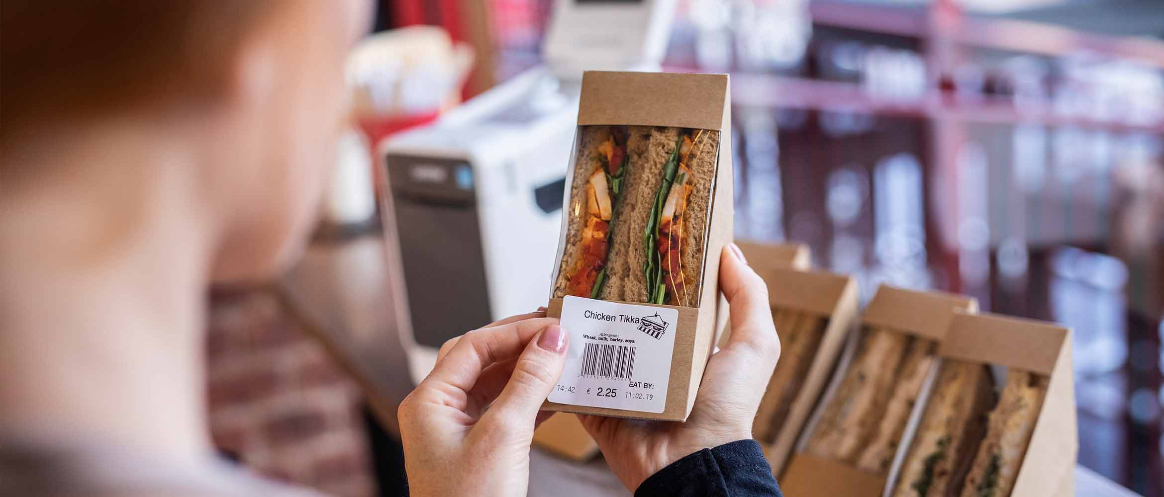 woman labelling a sandwich in a food retail environment with a Brother food safety TD labeller, showing allergen information in bold