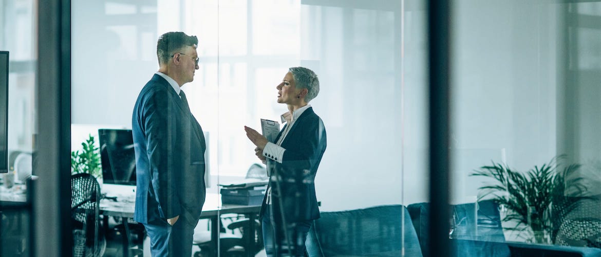 A business woman holding a clipboard and gesturing while explaining something to a business man in an office environment