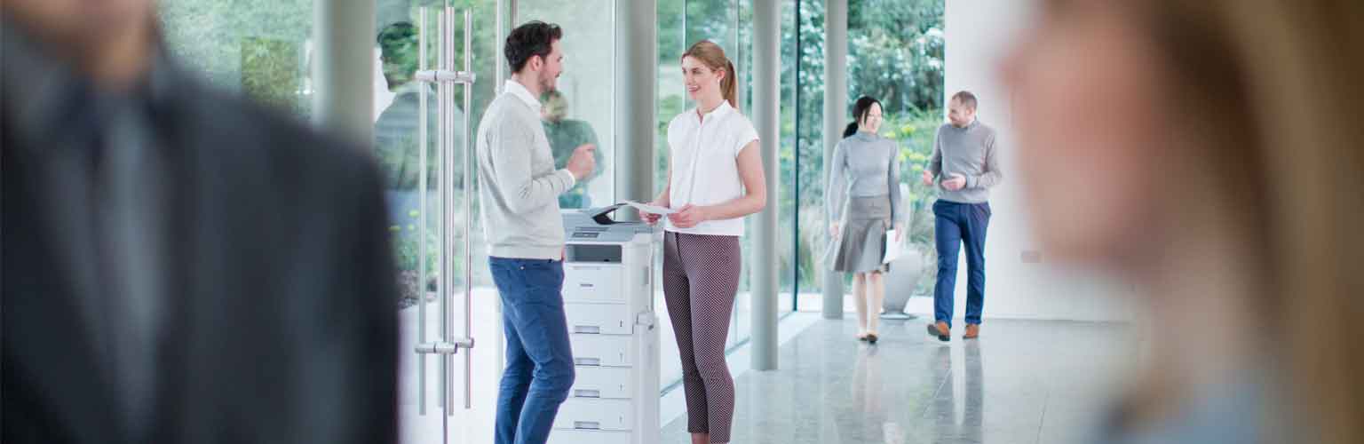 Two colleagues chatting at a Brother printer and two colleagues walking along in the background