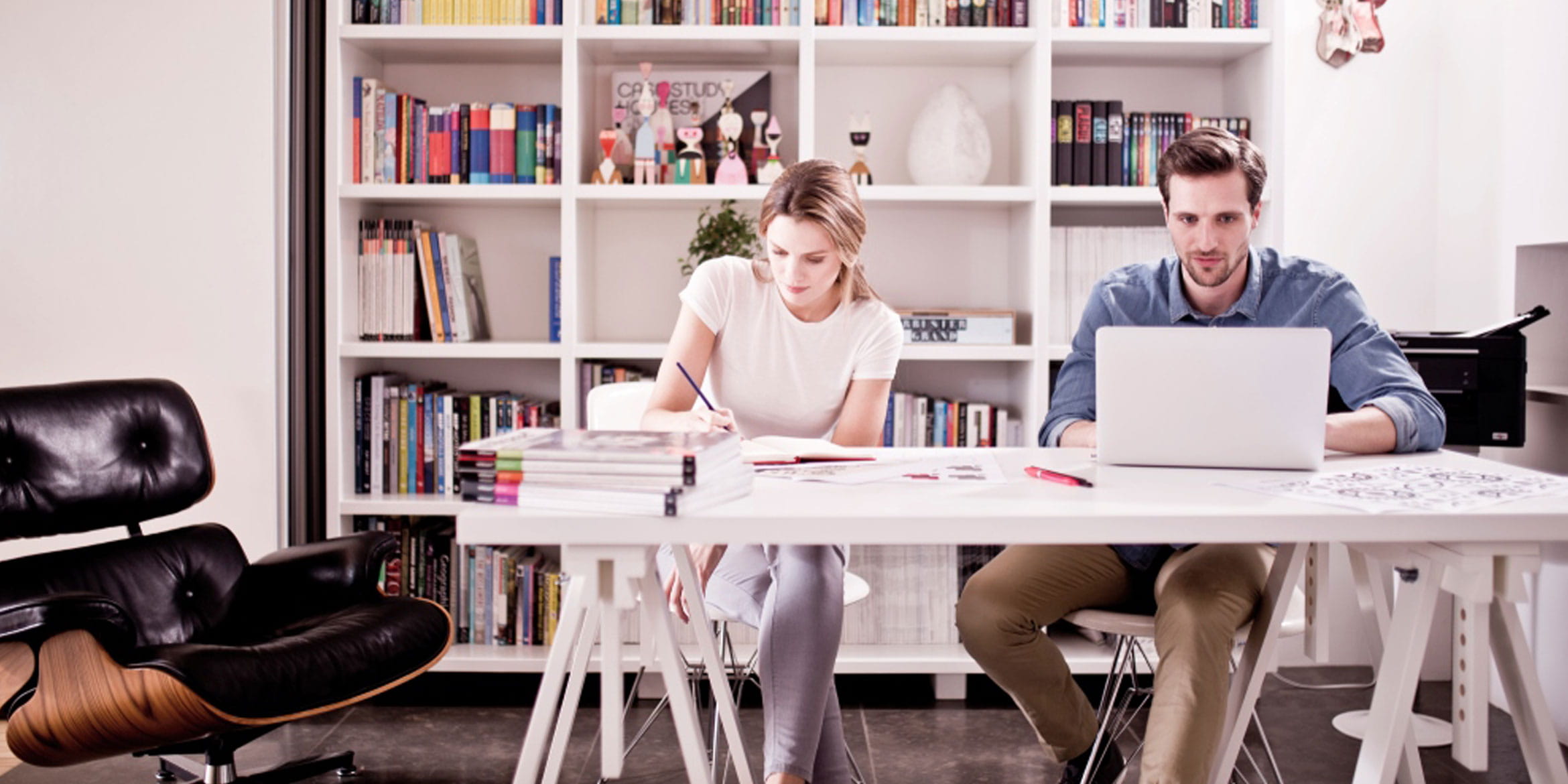 Woman writing and man using laptop computer at table with bookshelf and Brother printer in background