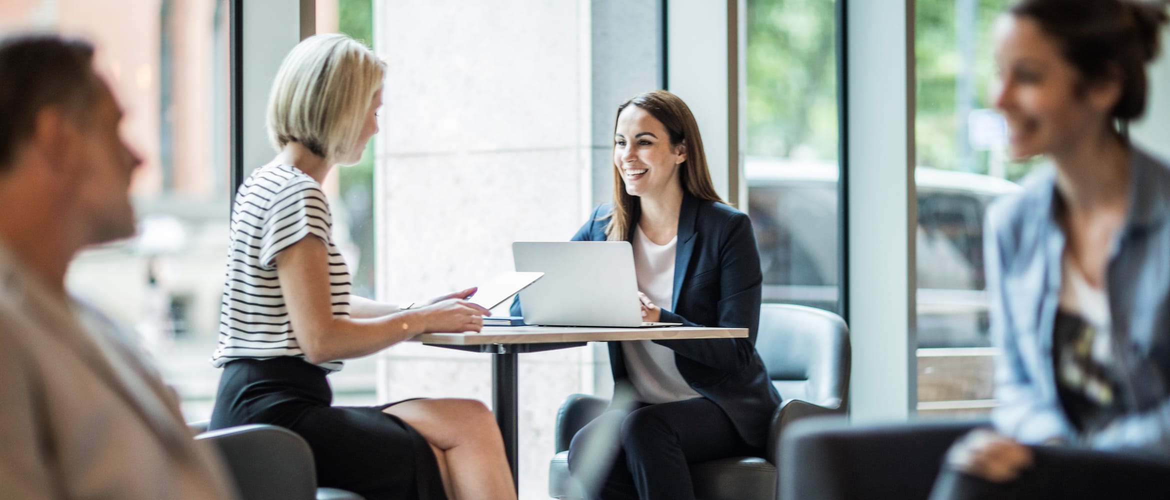 Two female work colleagues having a meeting at a coffee table with other people talking informally in the foreground