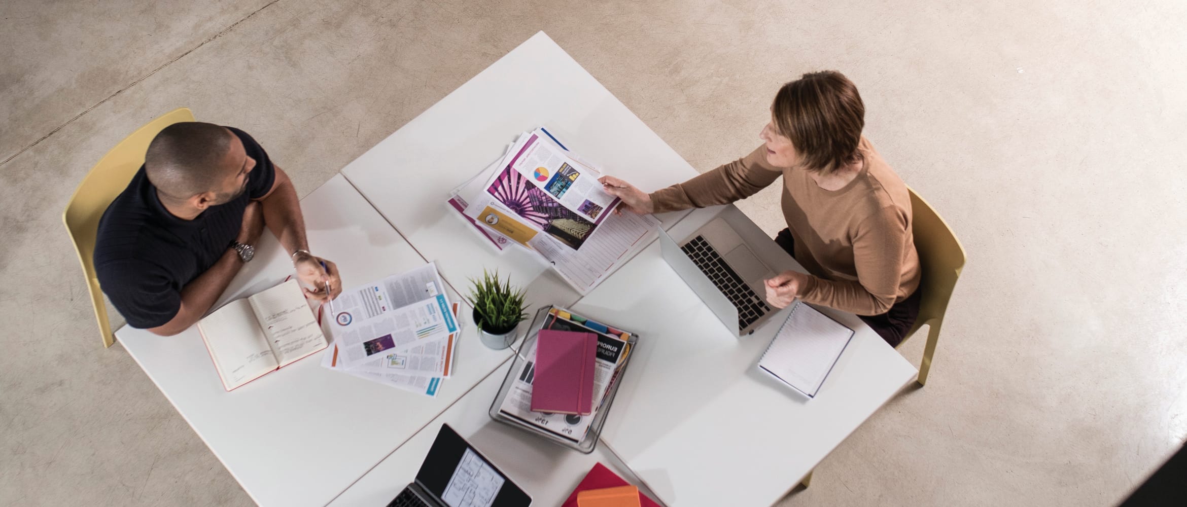 An overhead view of a man and woman working on a cluster of white tables with work items scattered over the surface