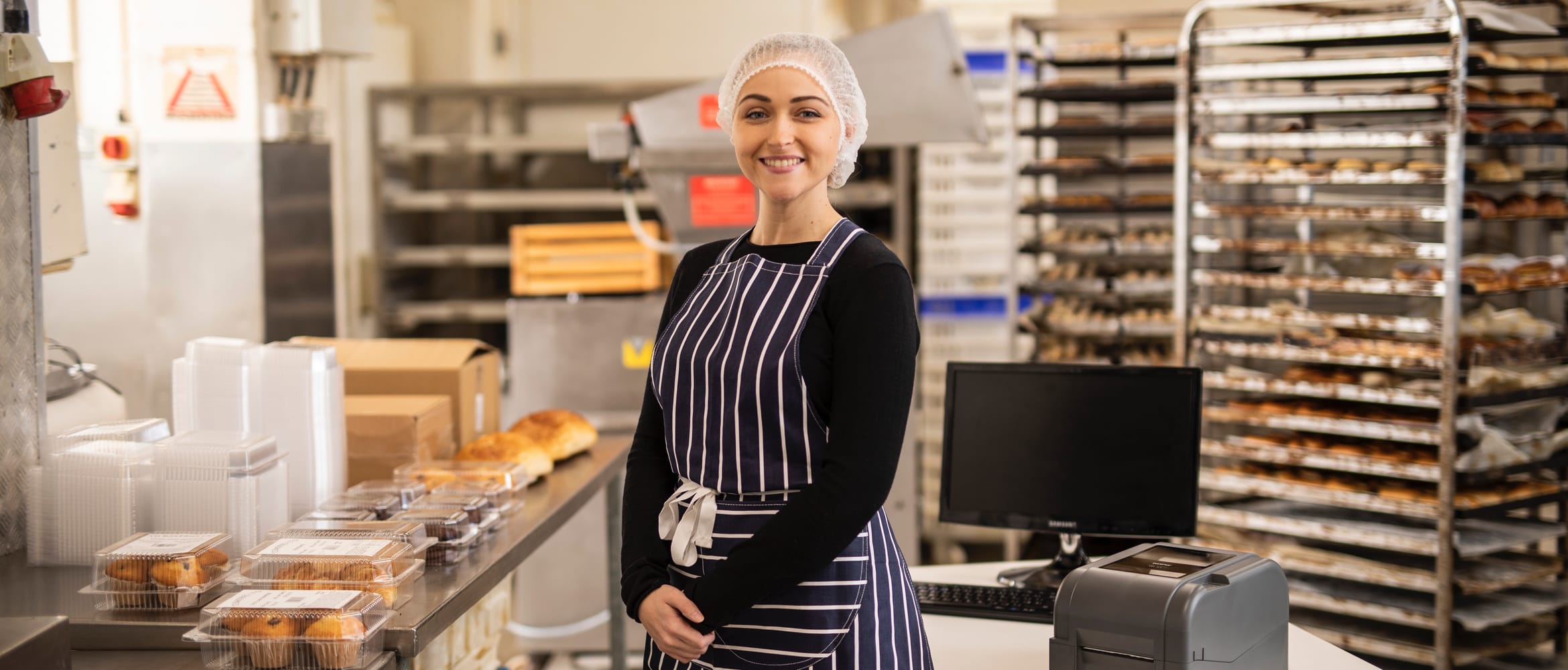 A female baker wearing a stripy navy apron smiling as she stands next to labelled food cartons on a counter in a commercial bakery kitchen