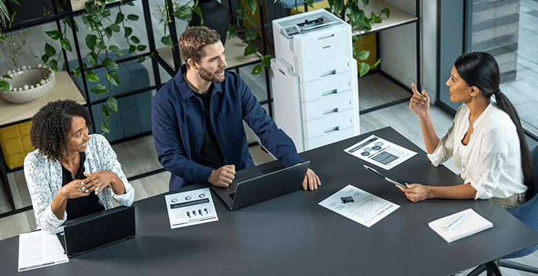 Three colleagues having a discussion around a table with a printer in the background in an SMB office environment