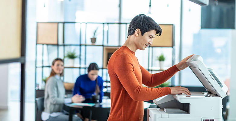 Man wearing orange jumper scanning document on Brother multifunction printer, 2 women sat at table in background