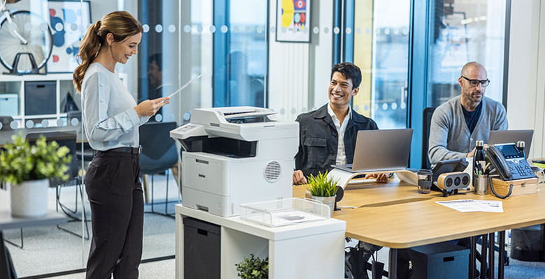 Busy office, 2 men sat working at desks, female stood at Brother printer holiding A4 document, plants, office furniture