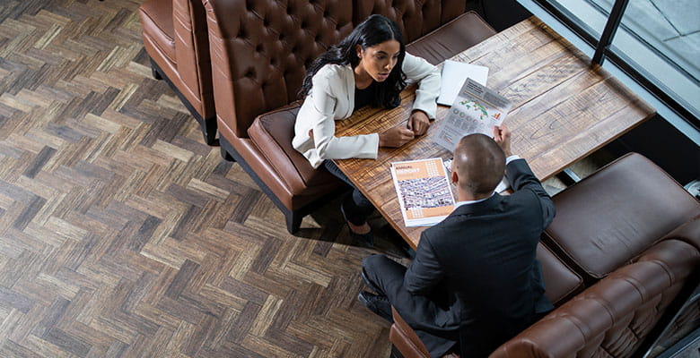 Man and woman sat across each other, A4 colour documents on wooden table