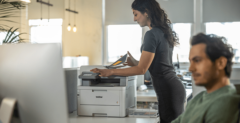 A woman printing a document in an office environment with a male colleague using a desktop computer in the foreground