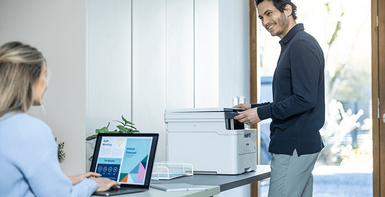 A man printing a document in an office environment with a female colleague using a laptop computer in the foreground