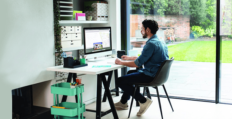 A man using an all-in-one computer while sat at a desk in front of sliding glass doors in a home office environment