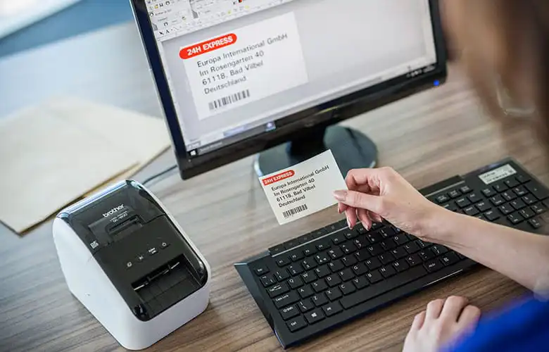 Brother QL printer next to monitor with person holding address label in office