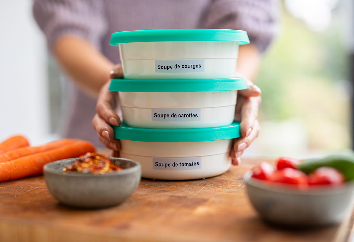 Femme tenant trois boîtes Tupperware étiquetées sur une planche à découper sur laquelle se trouvent des carottes, des tomates et des concombres.