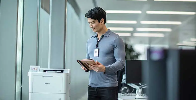 Man wearing lanyard holding tablet next to Brother printer in office