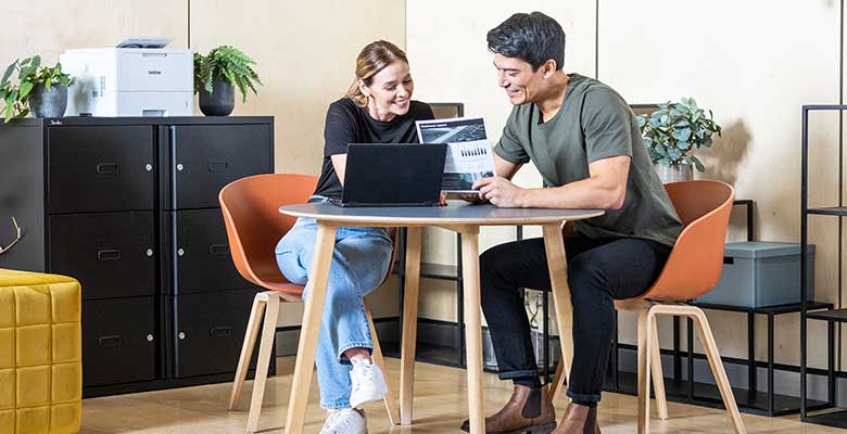 Man and woman sat at desk in office with printer, cabinets, plants